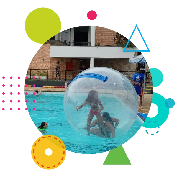 A imagem mostra um cenário de recreação aquática. No centro, duas crianças estão dentro de uma grande bola inflável transparente, que está flutuando na piscina. Uma das crianças está em pé, enquanto a outra está agachada. A piscina está localizada em um ambiente ao ar livre com uma área de observação ao fundo, onde alguns adultos estão presentes. Há também um instrutor ou supervisor vestido com um uniforme azul do SESC, que parece estar monitorando a atividade. A imagem é enquadrada em um círculo com várias formas geométricas coloridas, como círculos, triângulos e linhas pontilhadas, que adicionam um toque lúdico e vibrante à composição.