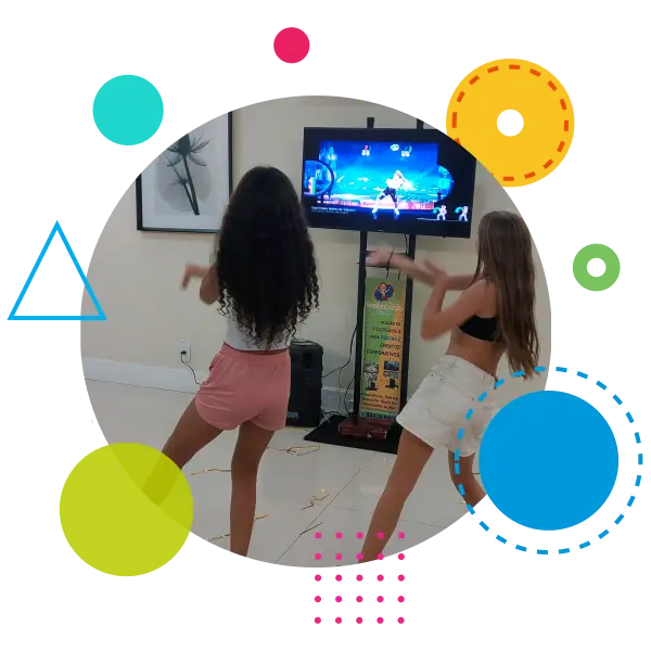 A imagem mostra duas meninas de costas, jogando um jogo de dança chamado Just Dance em um videogame. Elas estão seguindo os movimentos que aparecem na tela de uma televisão, que está montada em um suporte. As meninas parecem estar se divertindo, imitando os passos do jogo. A decoração ao redor inclui quadros na parede e um ambiente bem iluminado. A imagem é enquadrada em um círculo com várias formas geométricas coloridas, como círculos, triângulos e linhas pontilhadas, que adicionam um toque lúdico e vibrante à composição.