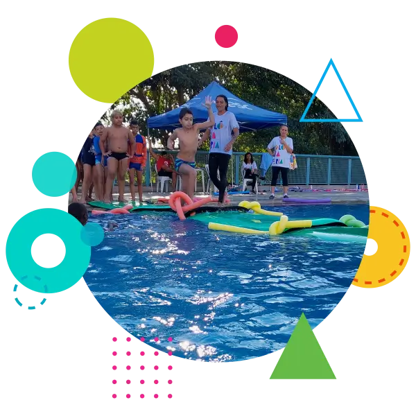 A imagem mostra uma cena de festa na piscina, com várias crianças e adultos participando de atividades aquáticas. No centro, um menino está prestes a pular na água de uma plataforma flutuante, rodeada de macarrões de piscina coloridos. Ao fundo, outras crianças e adultos observam a ação, alguns usando trajes de banho e outros com camisetas. Há uma tenda azul montada, indicando que o evento pode ser organizado e supervisionado. A piscina está reluzente sob a luz do sol, e a atmosfera é de diversão e descontração. A imagem é enquadrada em um círculo com várias formas geométricas coloridas, como círculos, triângulos e linhas pontilhadas, que adicionam um toque lúdico e vibrante à composição.